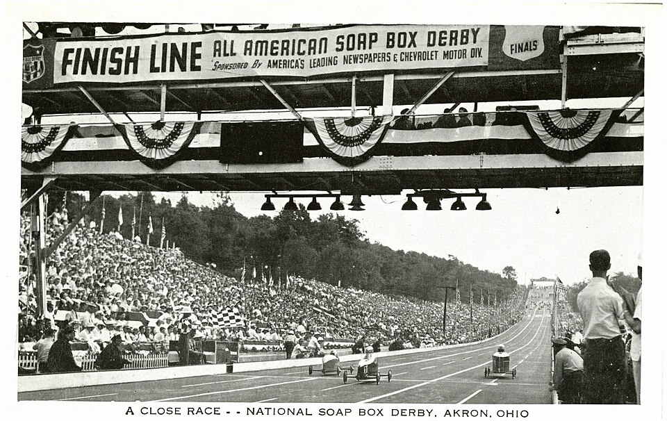 Three early Soap Box Derby cars racing toward the All-American finish structure as a packed hillside watches