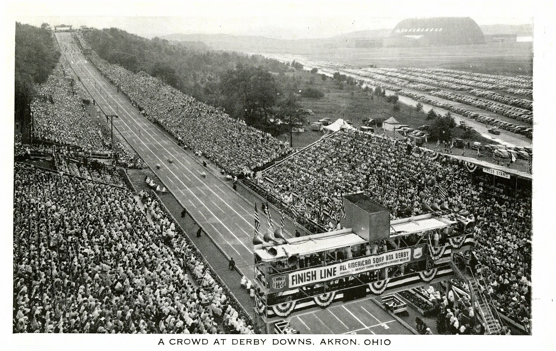 Aerial historic view of Derby Downs with spectators densely filling both hillsides, the finish line in front, and the Goodyear Airdock in the distance