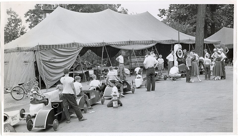 A long row of hand-built Soap Box Derby cars and racers waiting beside a large scale under inspection tents in 1953