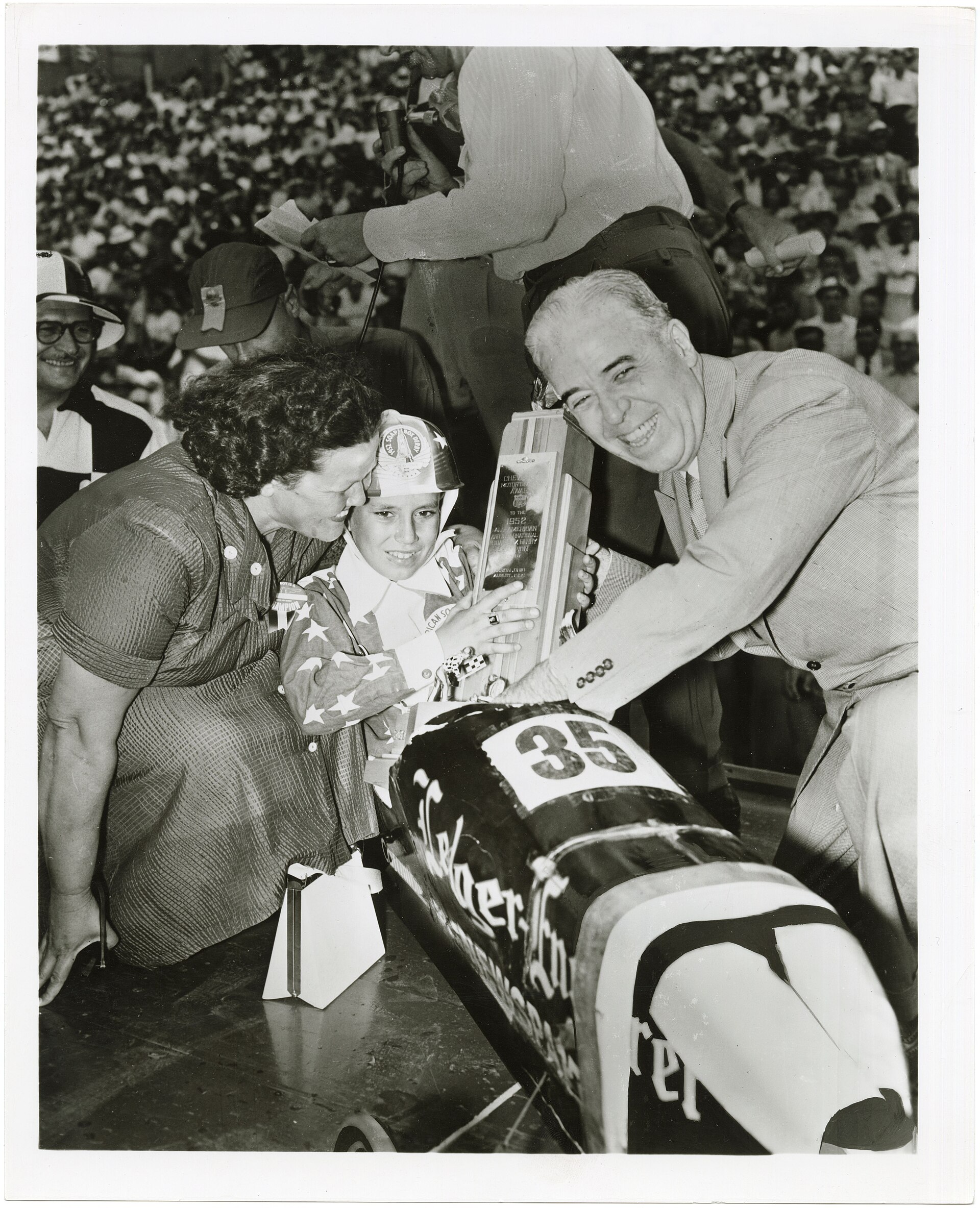 Joe Lunn holding the 1952 championship trophy beside his number 35 car, officials, photographers, and a crowd