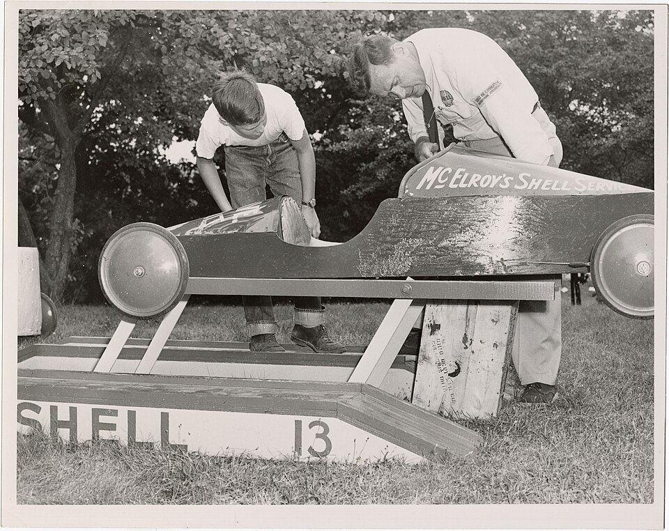 A young racer and a Shell representative inspect a long wooden Soap Box Derby car on a raised stand in 1953